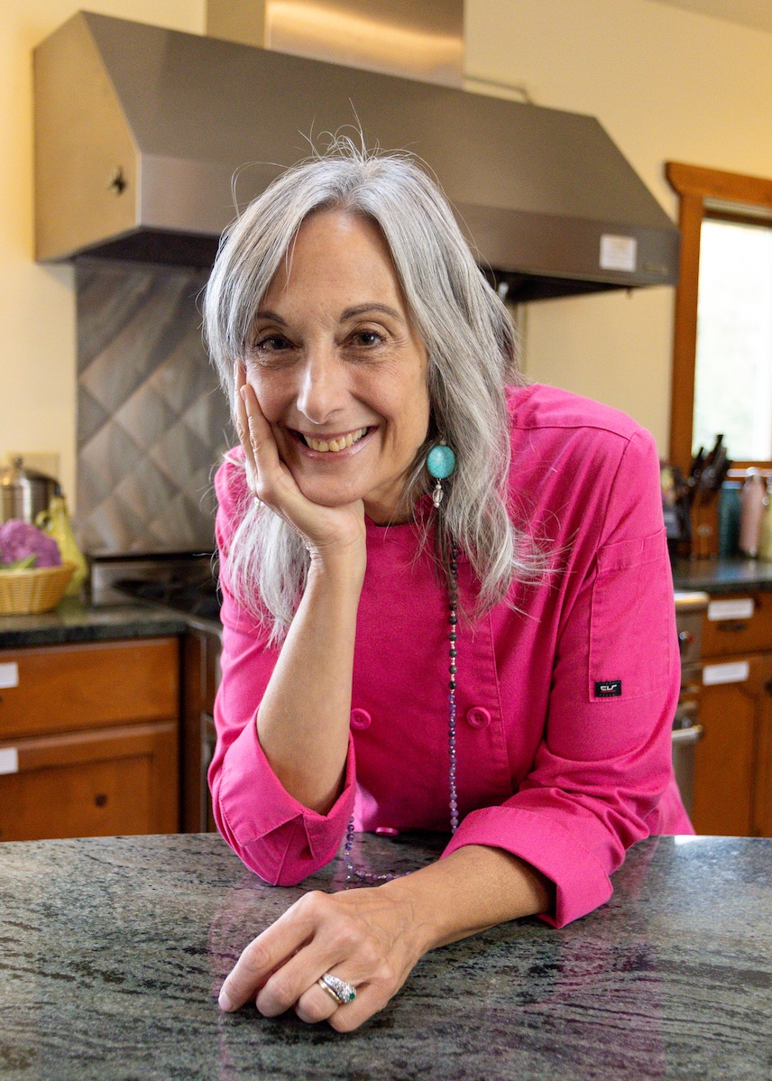 Cathy Vogt Personal Chef standing in a kitchen leaning on a marble countertop kitchen island in a pink chef jacket